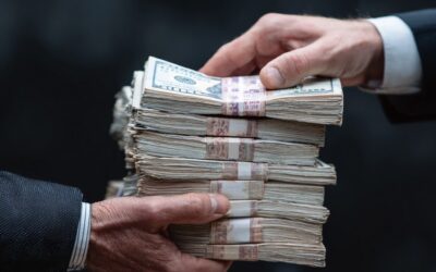 Two people in business attire exchange large stacks of U.S. hundred-dollar bills, held together with rubber bands, against a dark, blurred background.